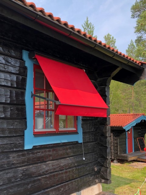 Klarröd fönstermarkis över fönster på timmrad stuga
